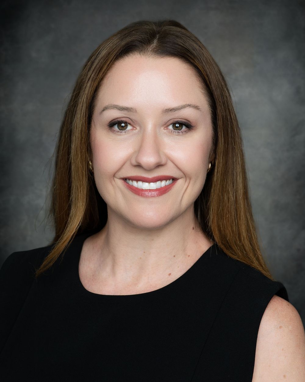 Professional headshot of a smiling woman with long brown hair wearing a black sleeveless top against a gray background.