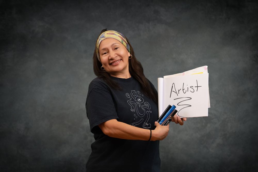 A smiling woman holds a white sign reading Artist, wearing a black t-shirt and headband against a gray background.