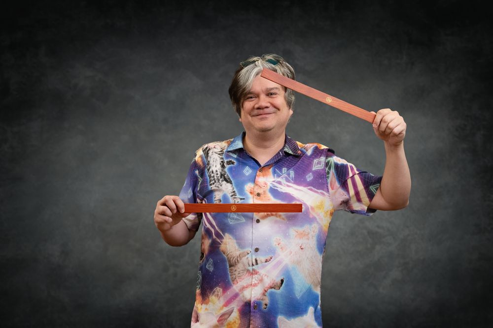 Man in colorful galaxy shirt holds a boomerang and wooden box against a dark gray background.