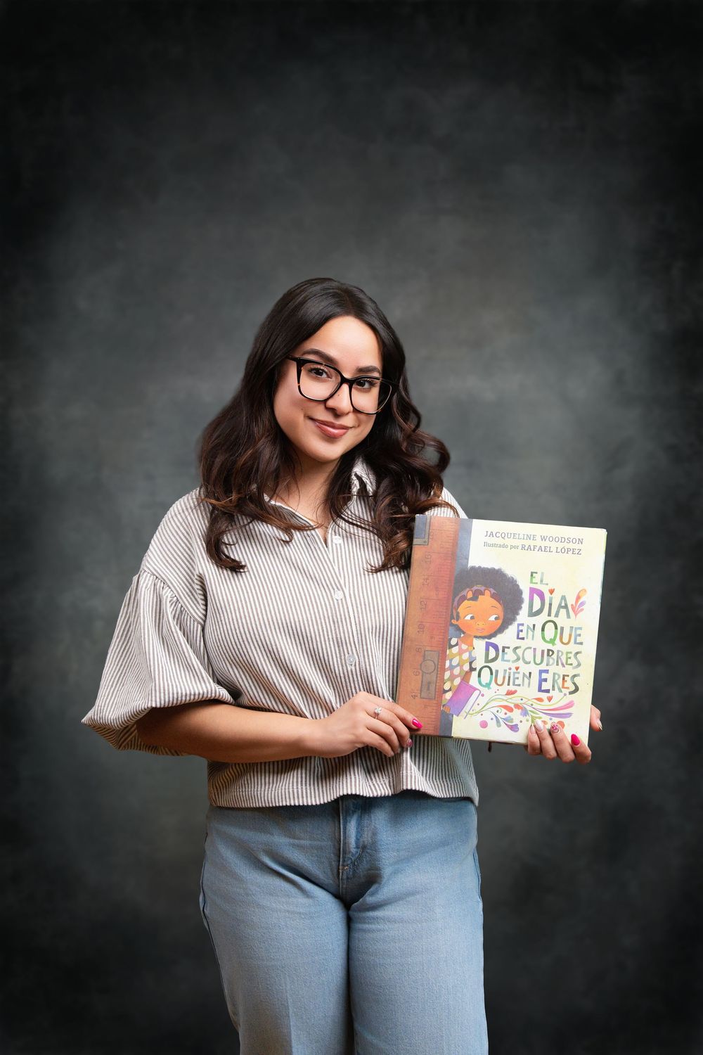 Young woman with glasses holding a Spanish children's book titled 'El Dia en Que Descubres Quien Eres' smiling.
