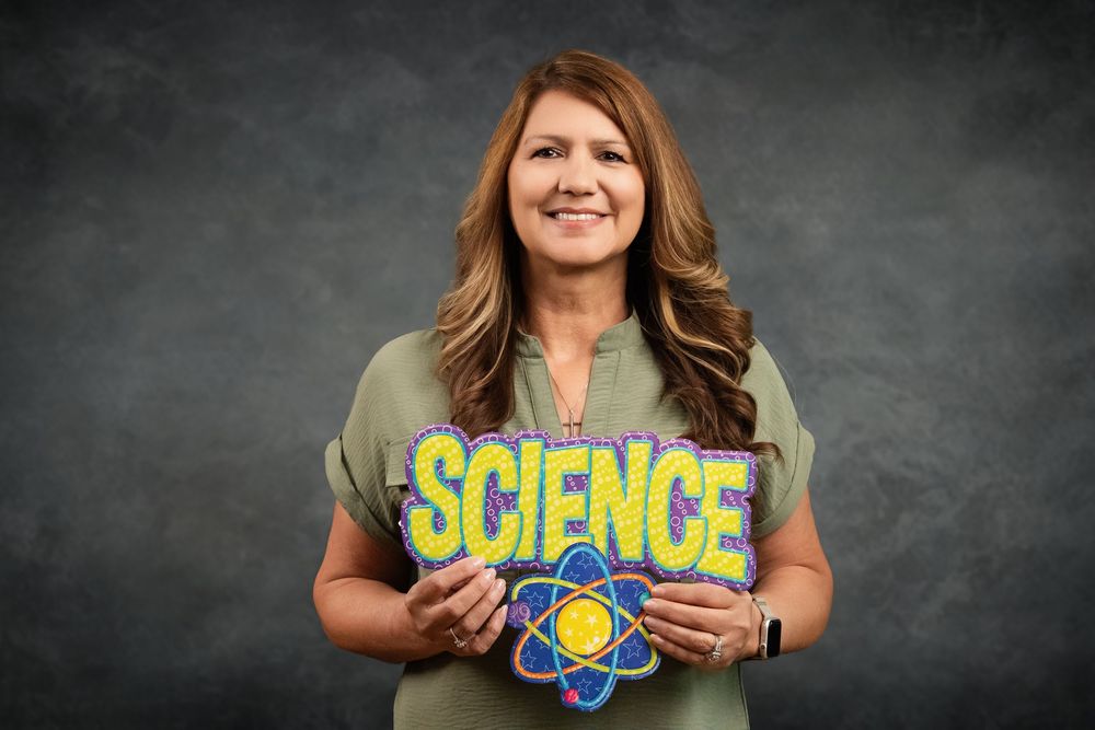 Smiling woman holding a colorful Science sign with atom graphic, posing against a gray background.