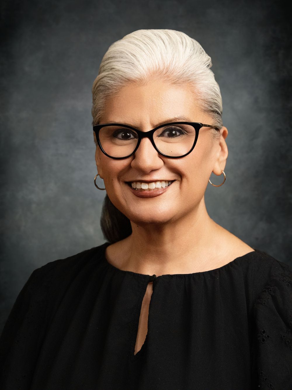 Professional headshot of a smiling woman with white hair, black glasses, and hoop earrings wearing a black top.