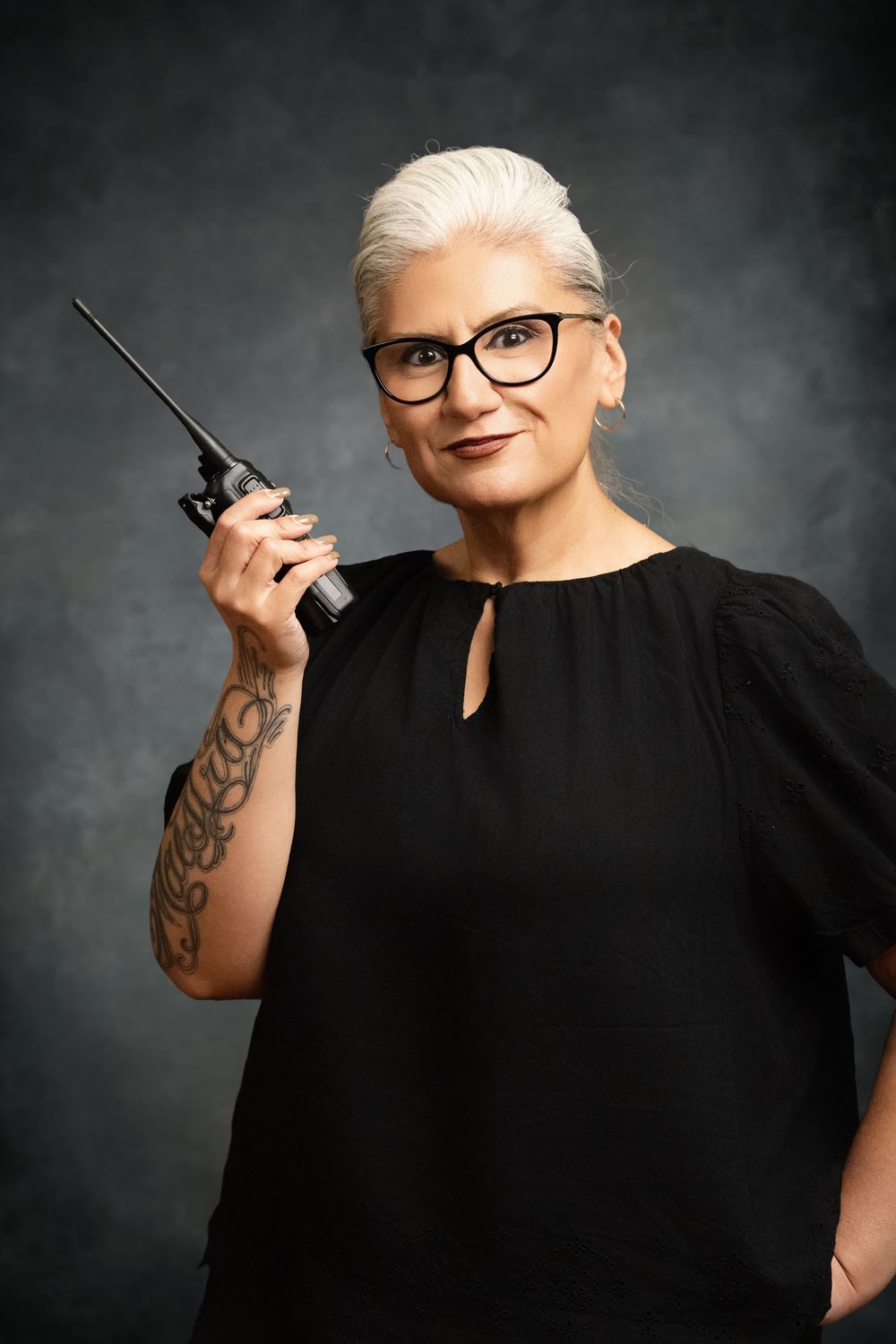 Mature woman with white hair and glasses holding a walkie-talkie, wearing black top with floral arm tattoo.
