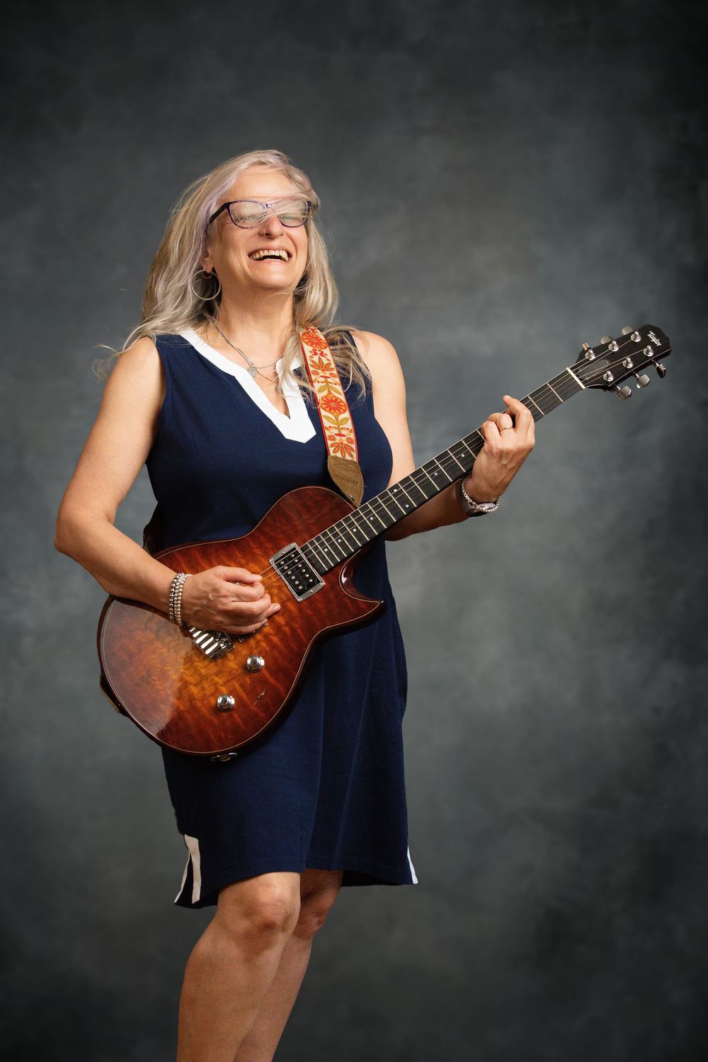 Smiling middle-aged woman with gray hair playing a brown electric guitar, wearing a navy dress and glasses in a studio.