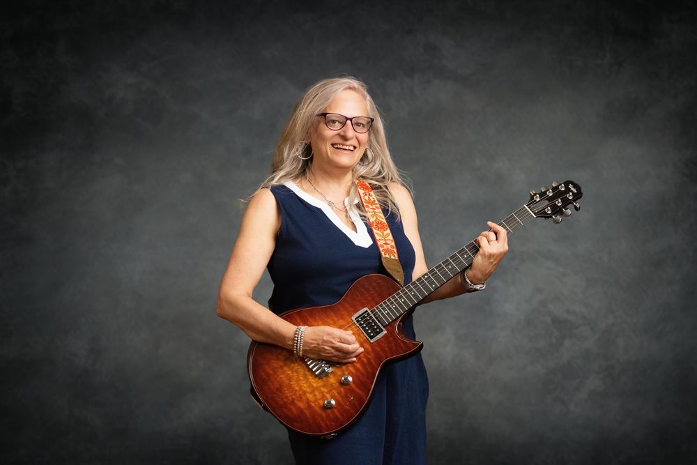 Smiling woman in navy dress playing a sunburst electric guitar against a gray studio background.