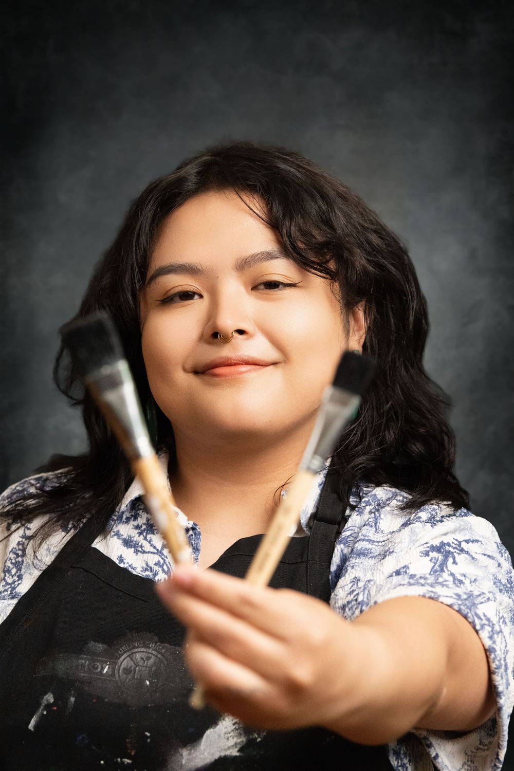 Young female artist in black apron holding paint brushes toward camera, smiling confidently against dark background.