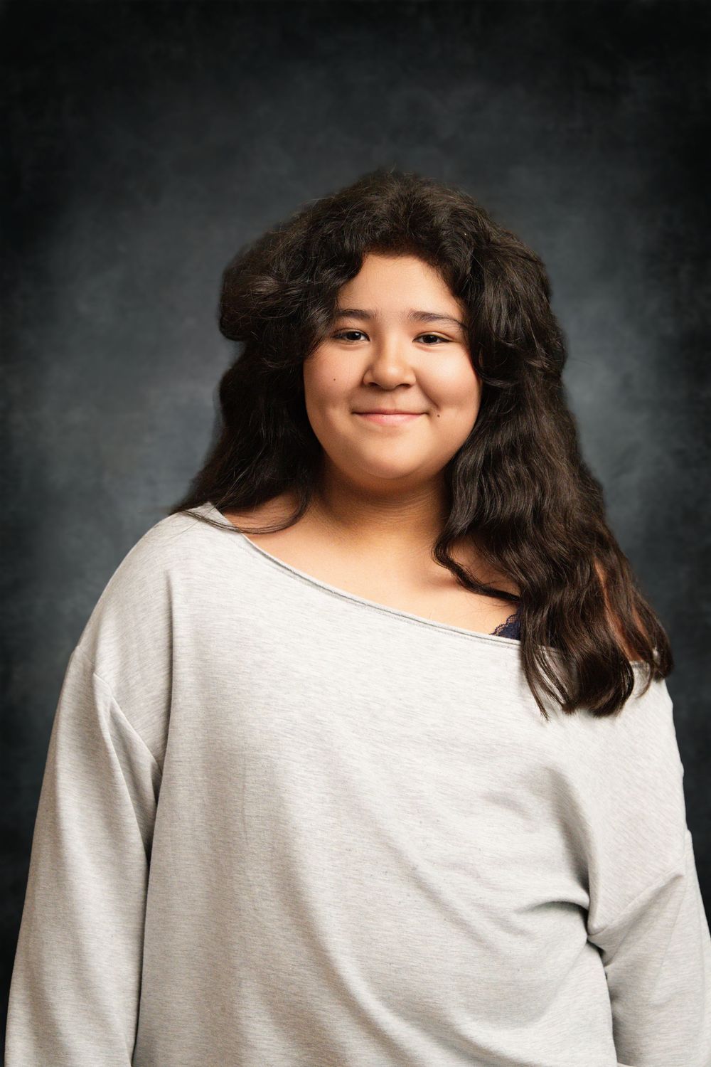 Young woman with long dark hair smiling, wearing a gray off-shoulder top, posed against a dark studio background.
