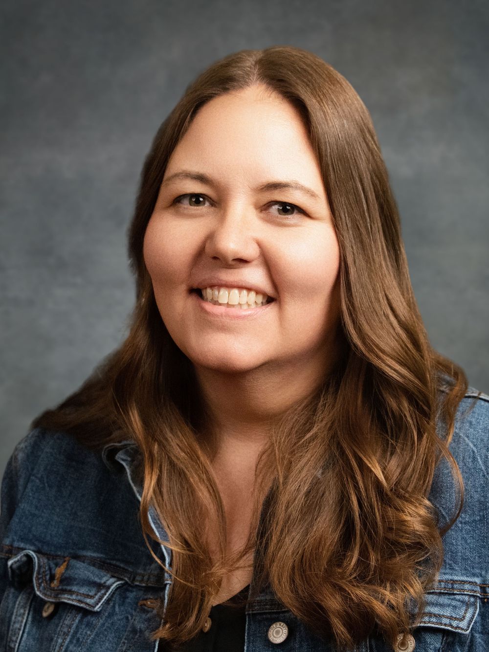 Smiling woman with long brown hair wearing a denim jacket poses for a professional headshot photo.