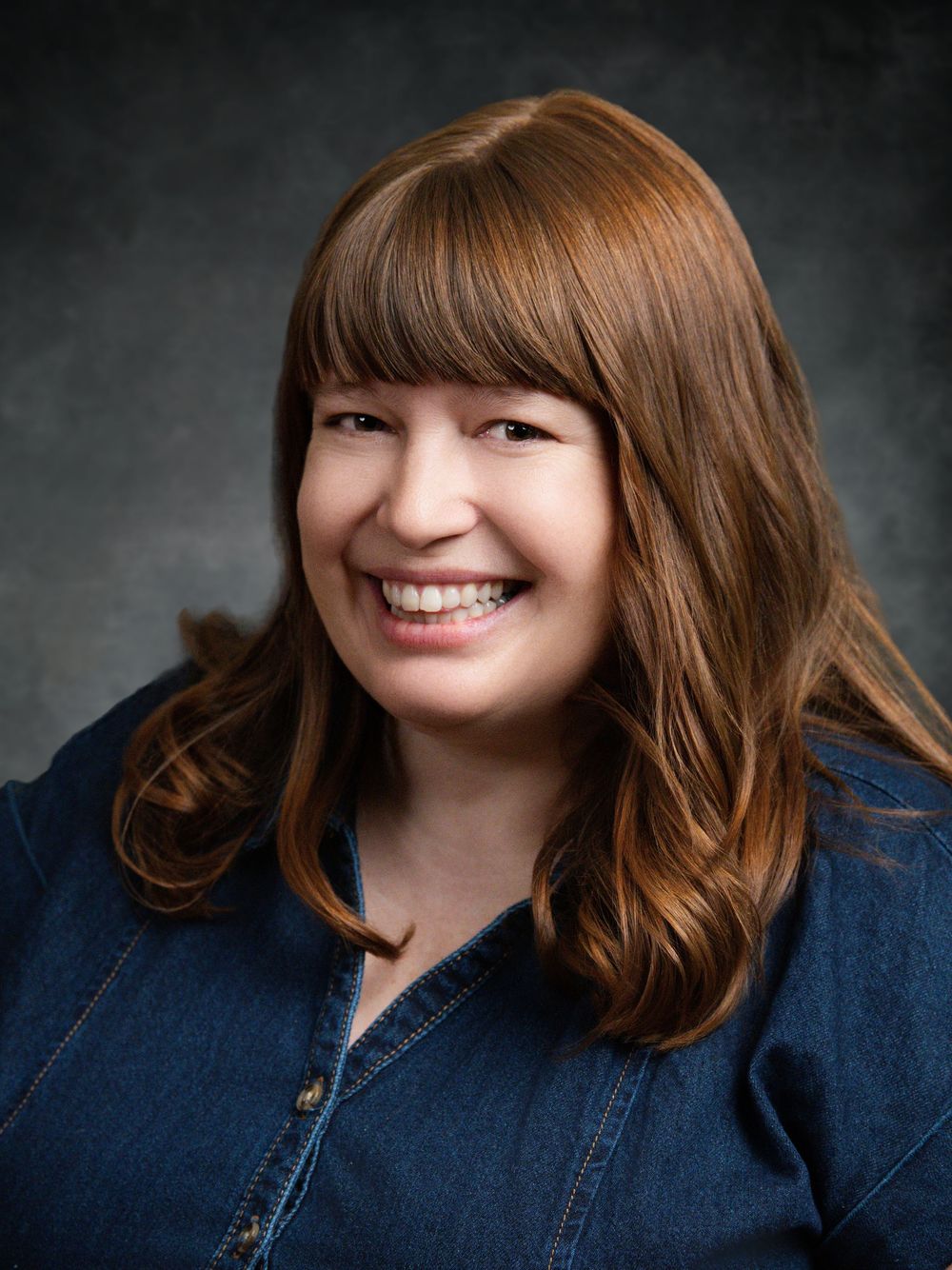 Smiling woman with red hair and bangs wearing a denim shirt against a gray studio background.