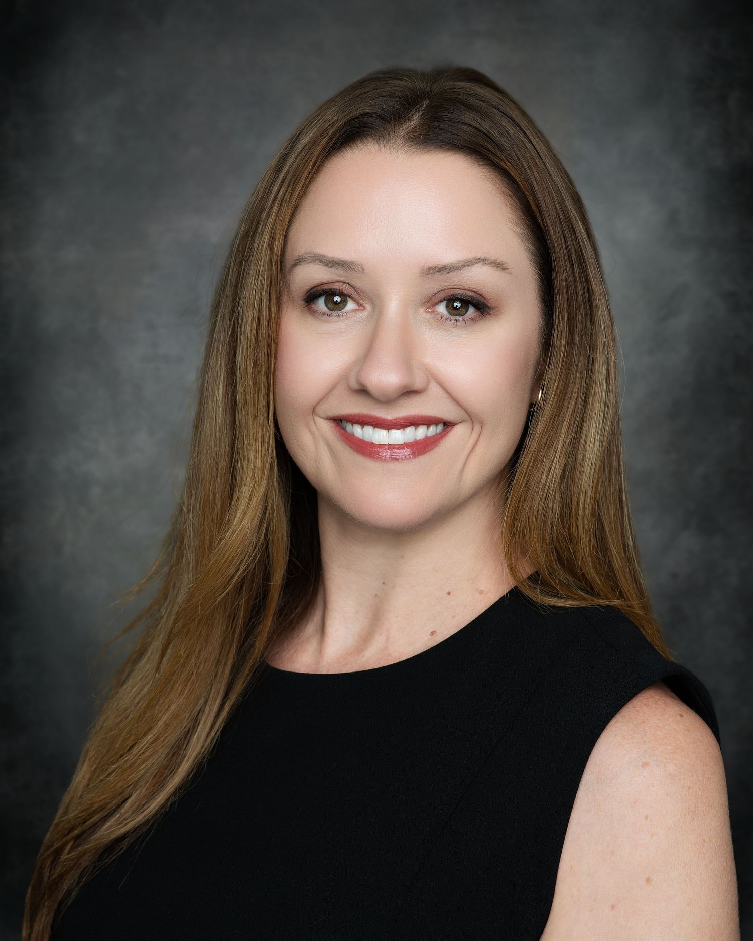Professional headshot of a smiling woman with long brown hair wearing a black sleeveless top against a gray background.