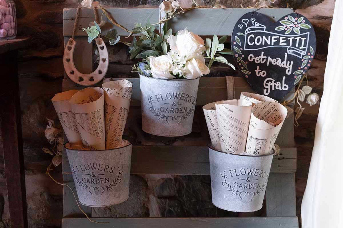 Rustic wedding confetti station with galvanized metal buckets holding paper cones and flowers on a wooden shelf.