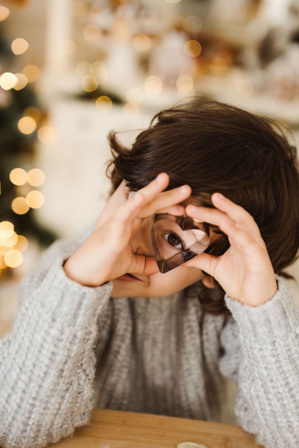 Child peeks through heart-shaped cookie cutter with Christmas tree lights bokeh background in cozy holiday kitchen setting.