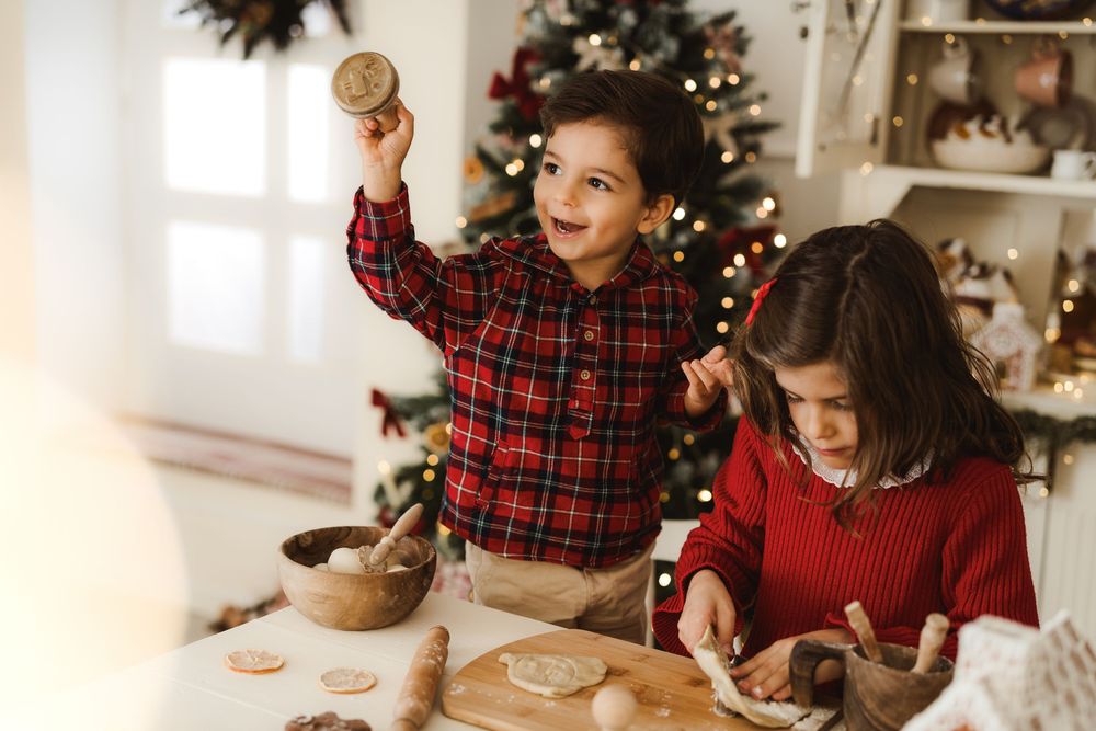 Two children in red holiday outfits bake Christmas cookies near a decorated tree.