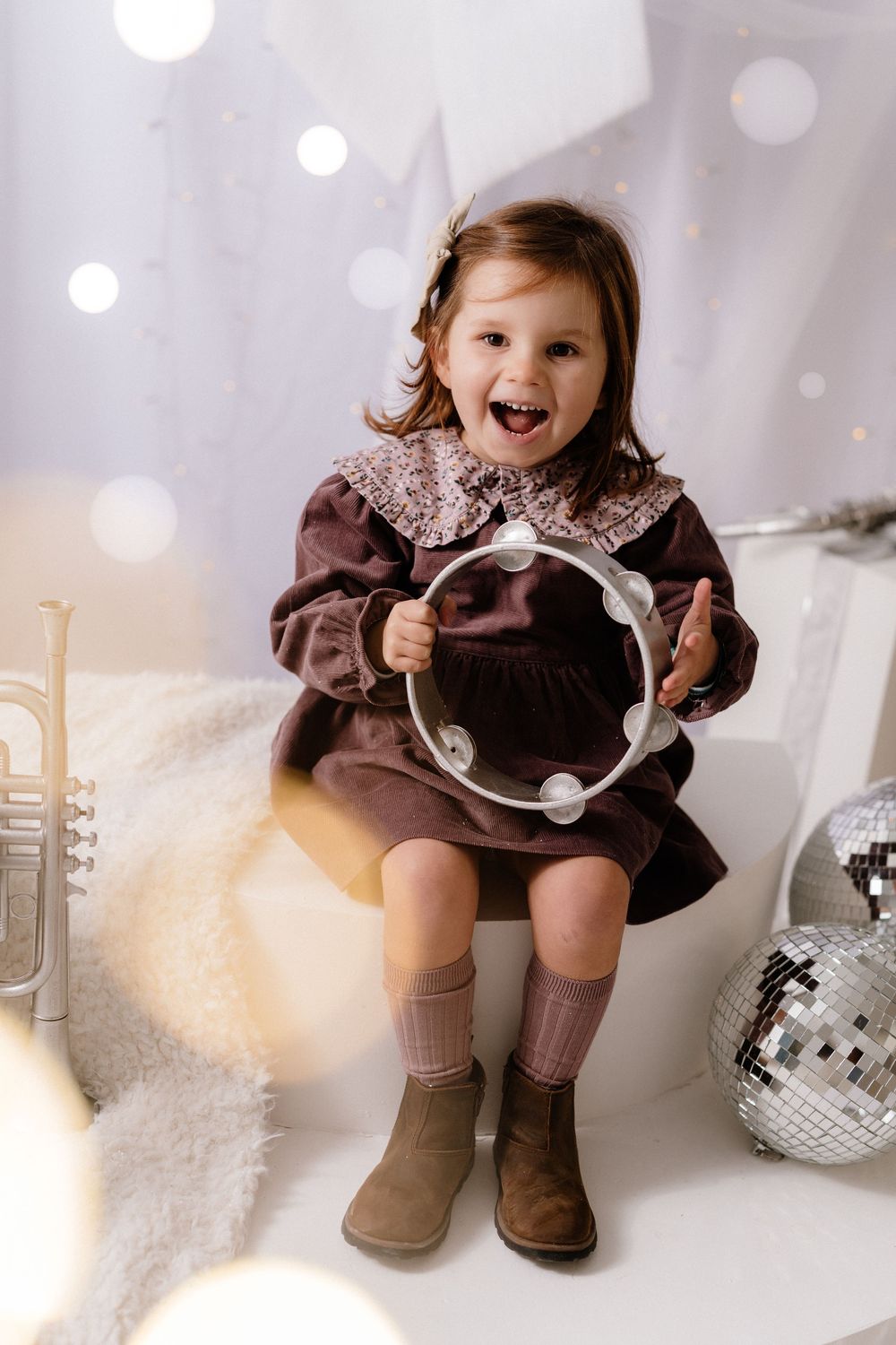 A joyful toddler girl in a brown dress holds a tambourine, surrounded by disco balls and a trumpet in a festive setting.