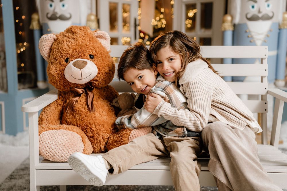 Two children hugging a large brown teddy bear on a white bench with festive holiday decorations in the background.