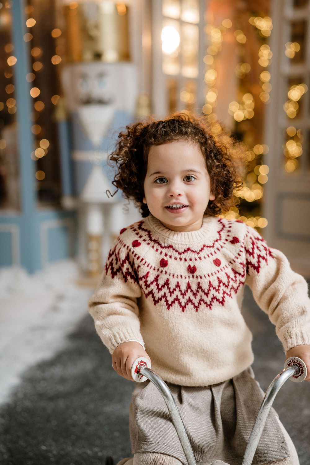 Curly-haired toddler in a Nordic sweater smiling while riding a balance bike near Christmas decorations.