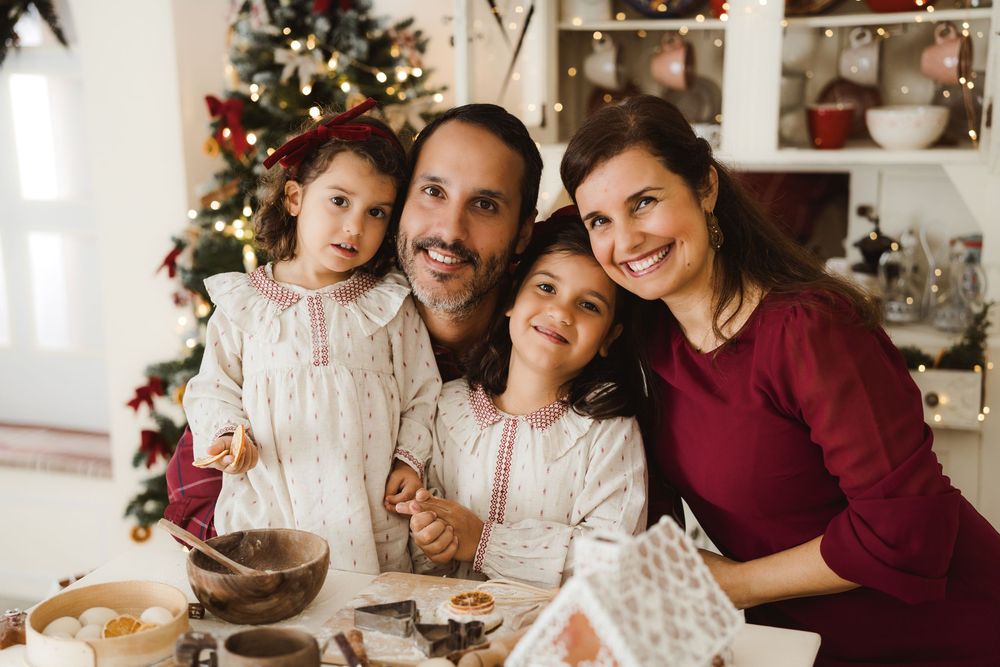 Family of four smiling together in festive kitchen with Christmas tree, baking holiday cookies and gingerbread house.