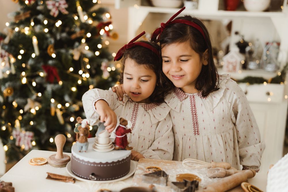 Two young girls in matching dresses decorating a chocolate Christmas cake near a decorated tree.