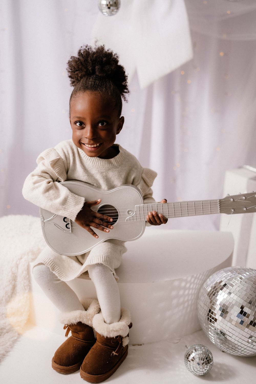 A smiling young girl in cream outfit and brown boots plays a white toy guitar near disco balls in a festive white setting.