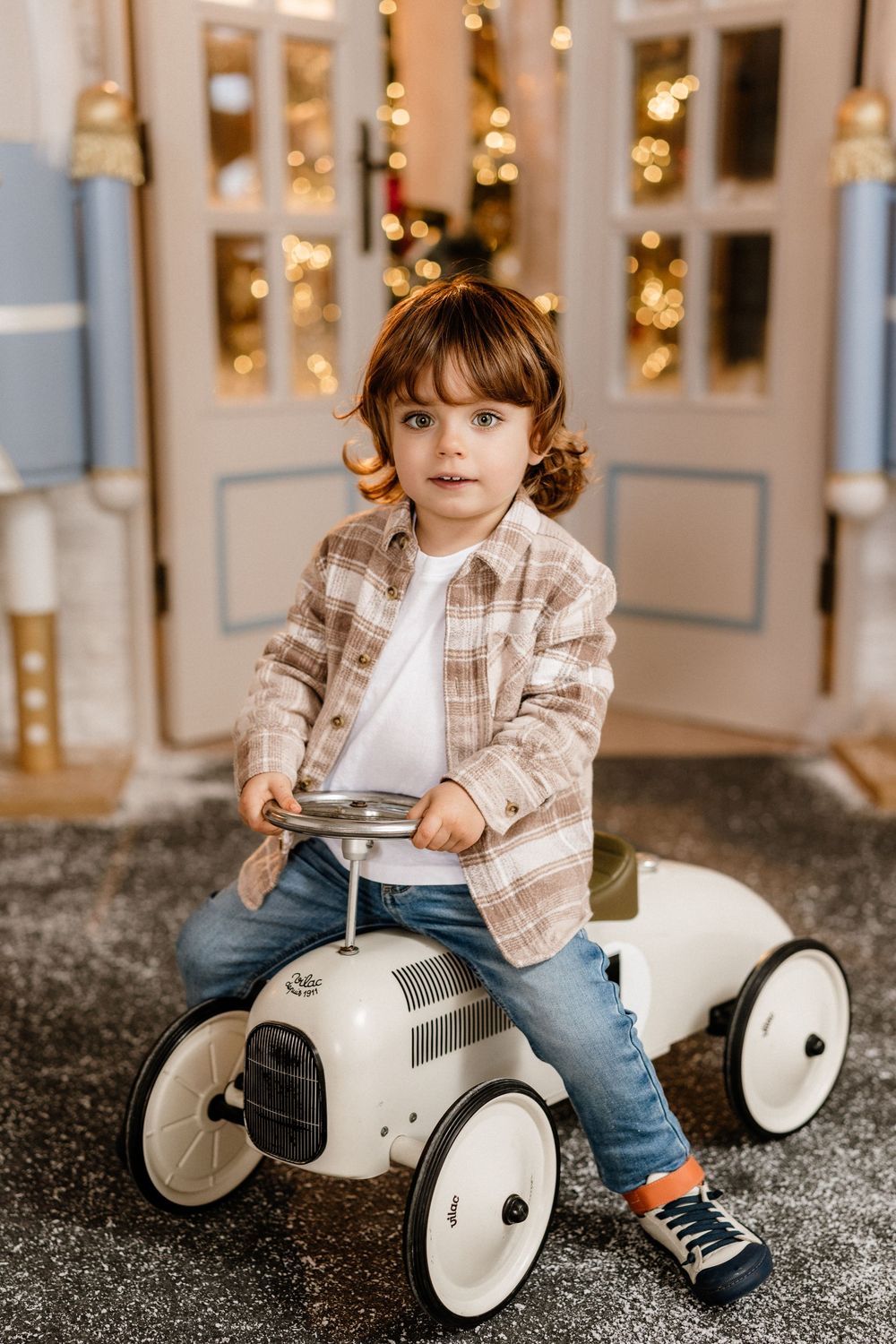 Young toddler boy riding a white vintage toy car in a festive room with bokeh lights in the background.