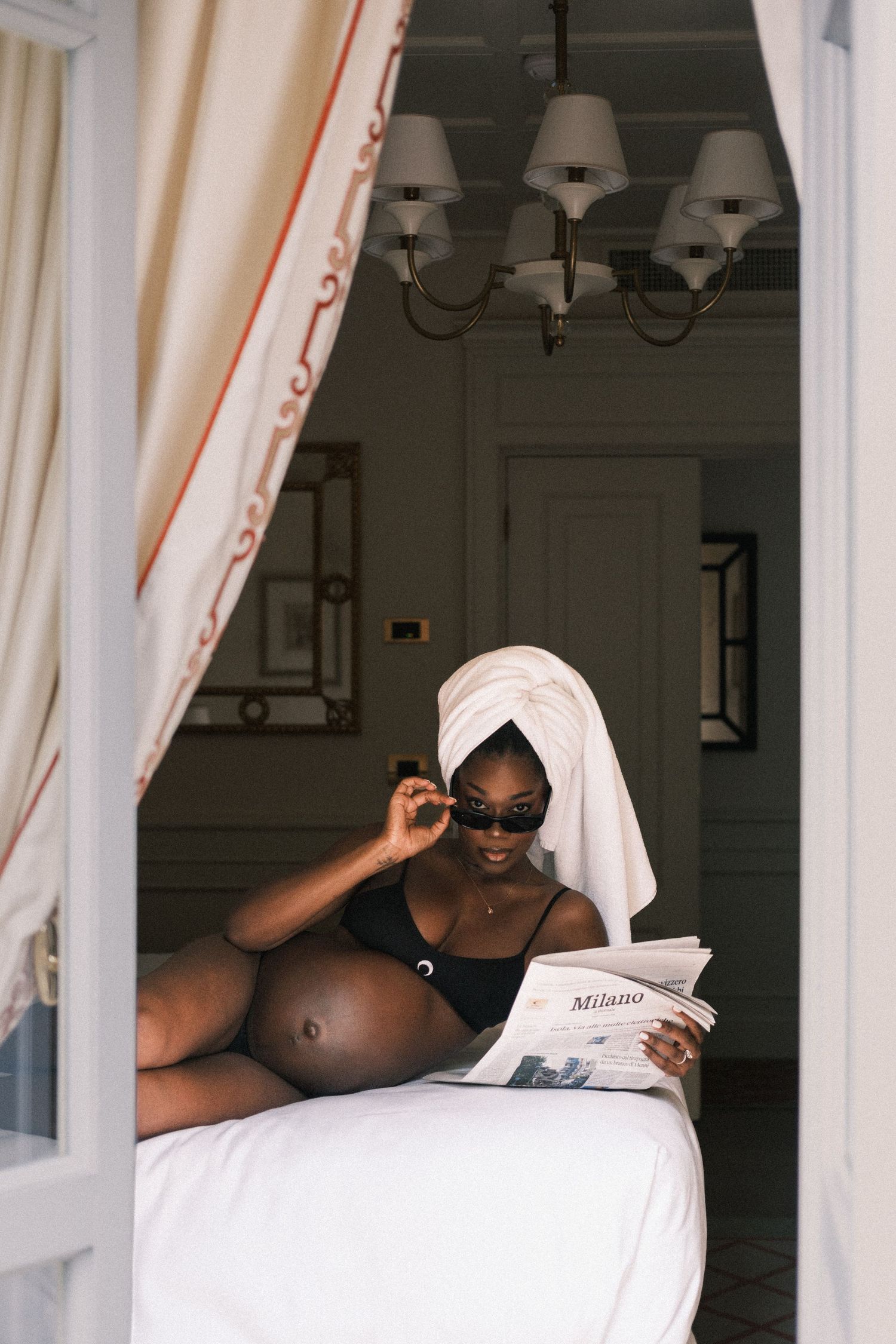 Pregnant Black woman in black bikini with towel turban reads Milano newspaper on white bed in elegant hotel room.