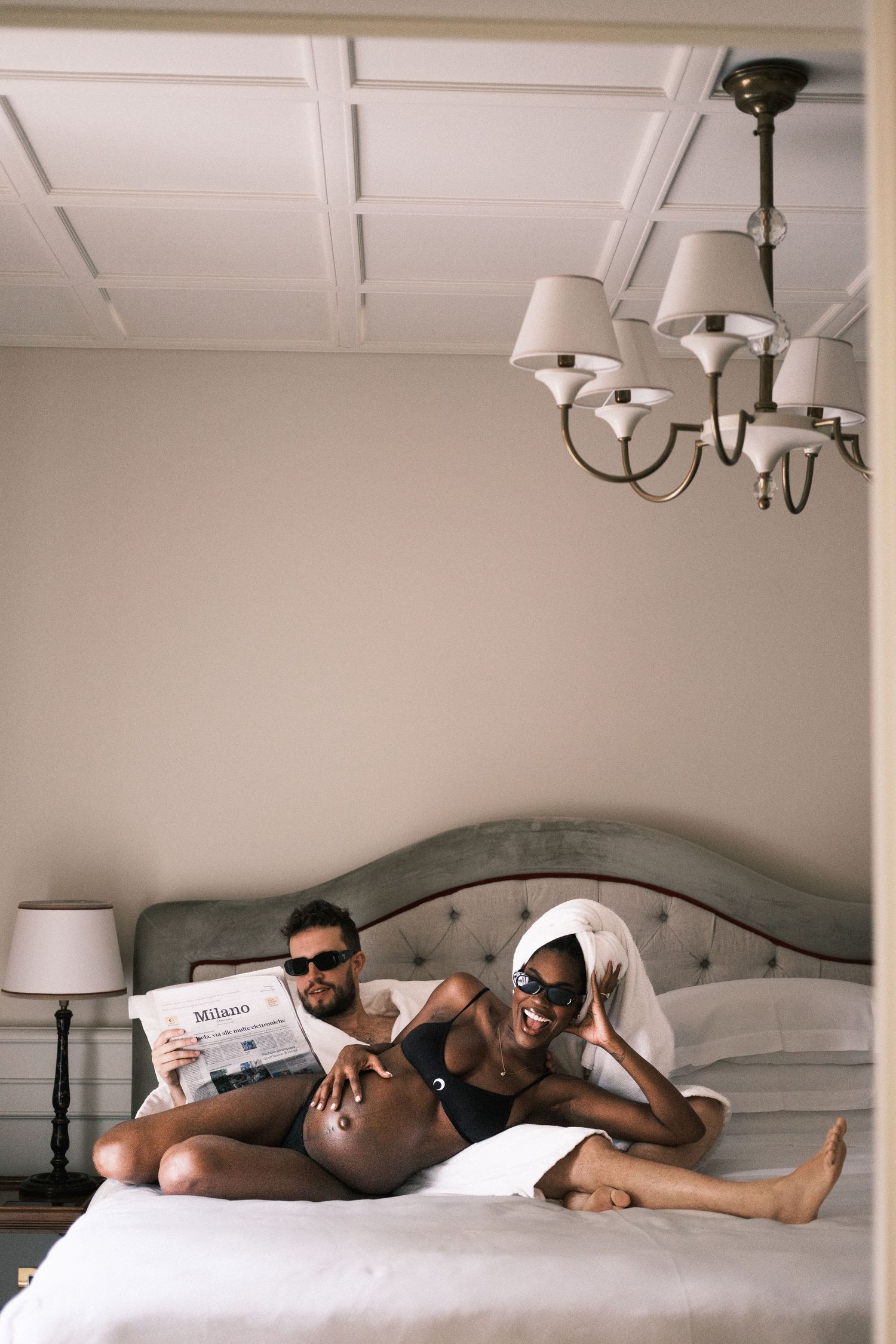 Couple relaxing on bed in elegant bedroom, man reading Milano magazine, woman in towel turban, both wearing sunglasses.