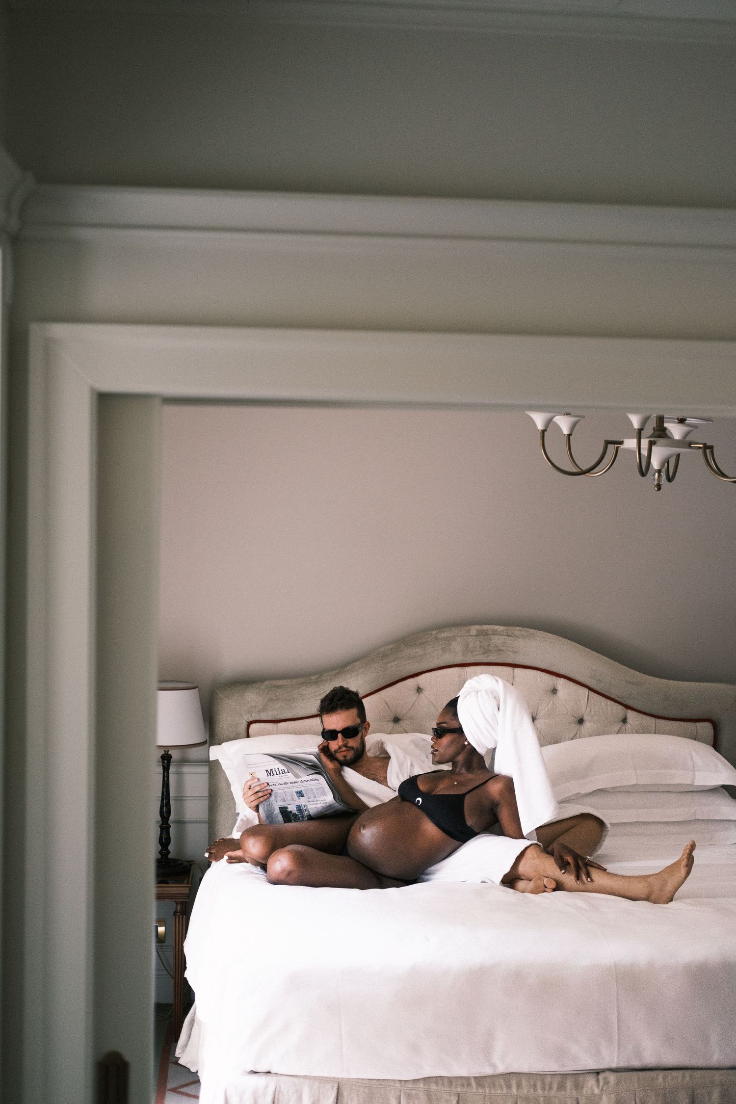Couple relaxing on a luxury hotel bed, man reading newspaper wearing sunglasses, woman with towel on head.
