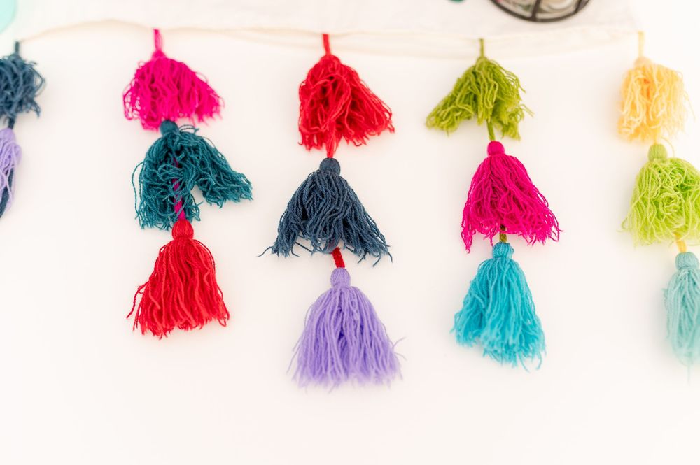 Colorful handmade yarn tassels in red, teal, navy, green, purple, and blue hanging on a white wall.