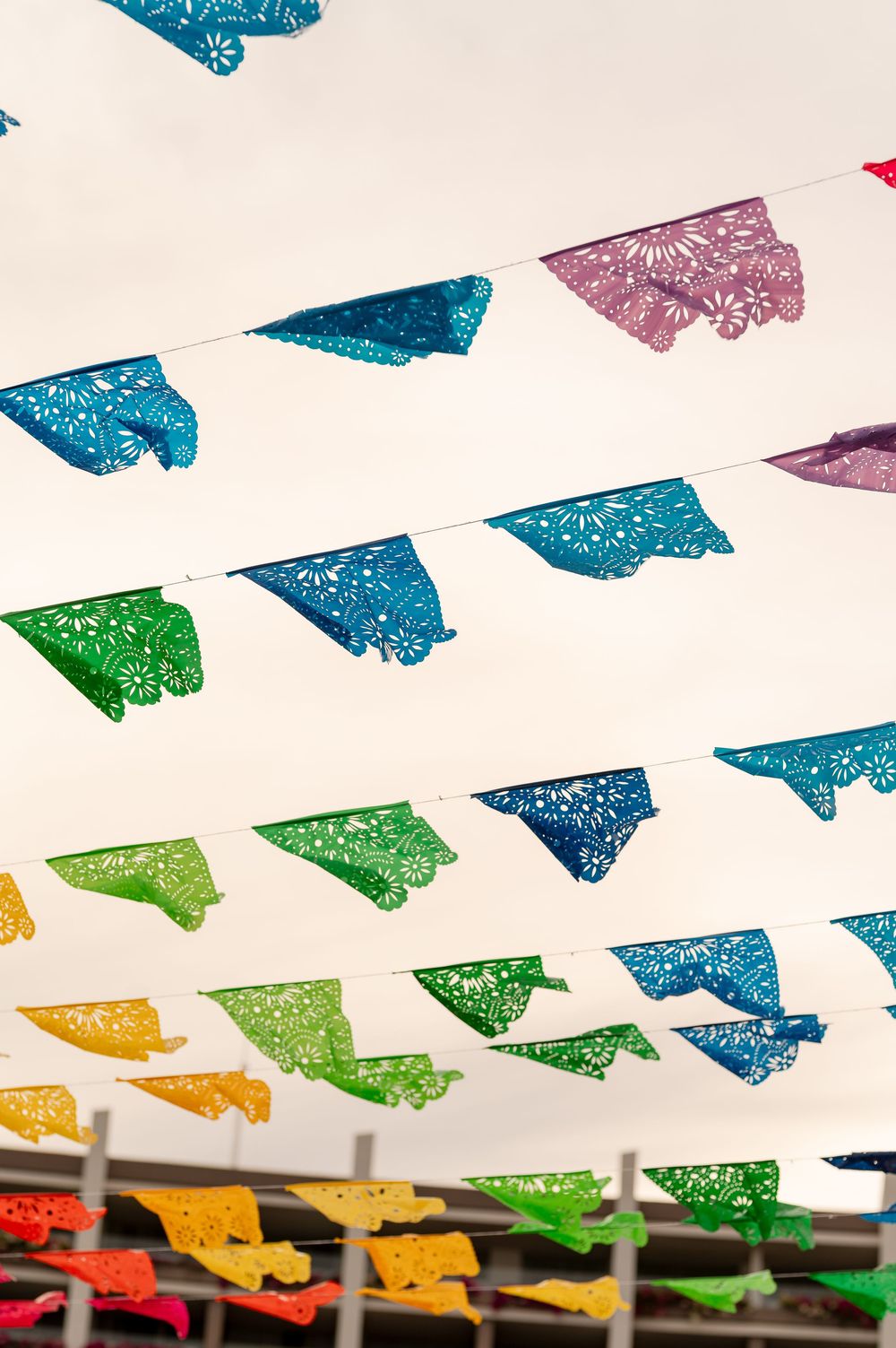 Colorful papel picado banners with intricate cut designs hanging on strings against a white sky.