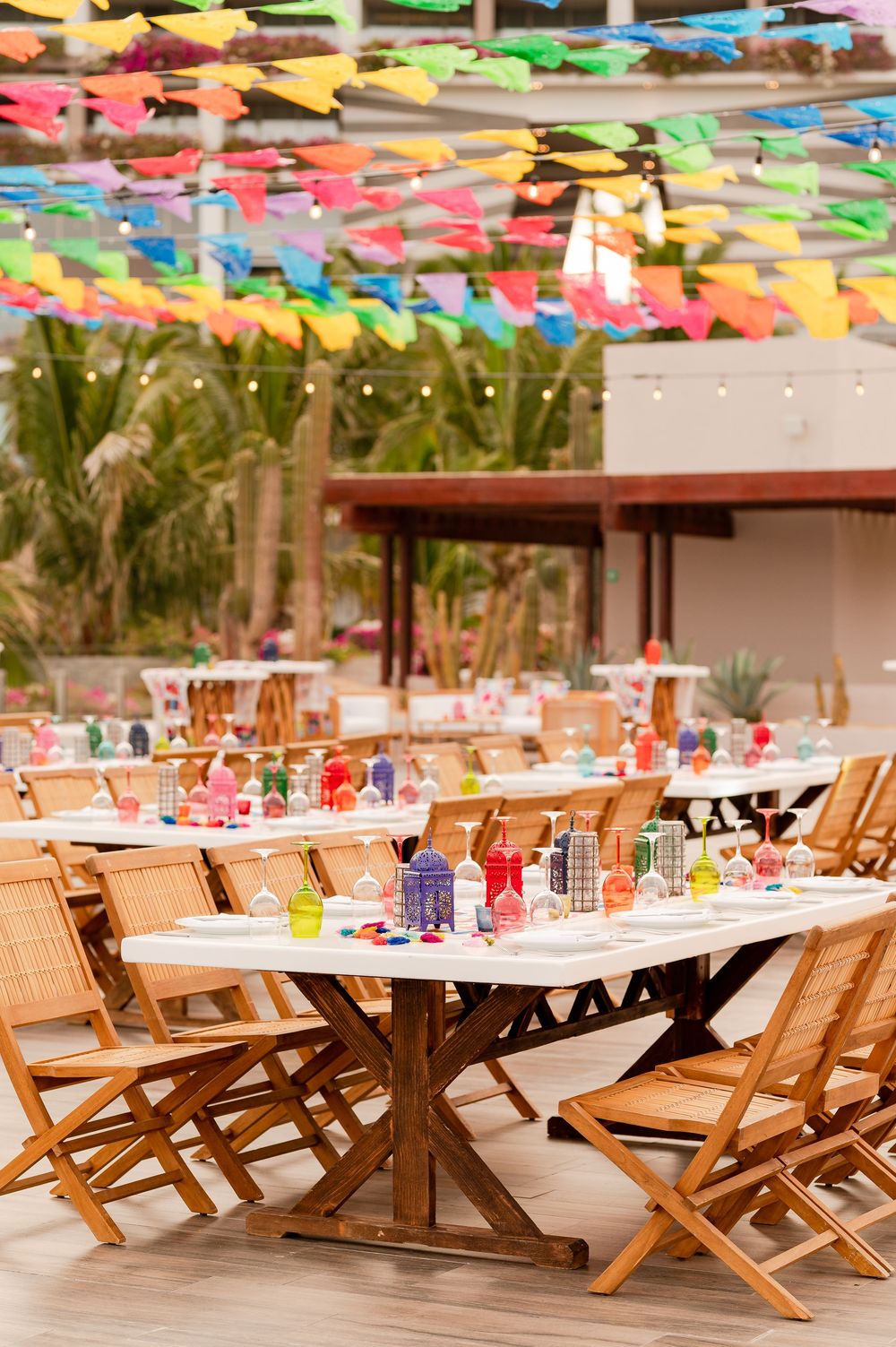 Colorful fiesta wedding reception with wooden chairs, white tables, vibrant decor, and rainbow papel picado banners overhead.
