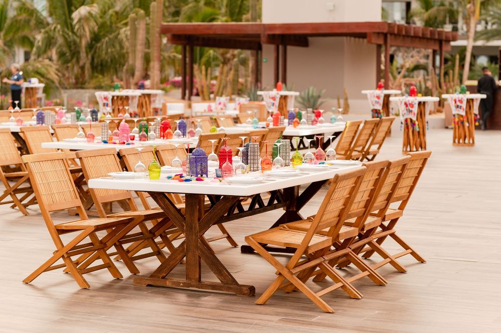 Outdoor dining area with wooden folding chairs and long tables set with colorful decor near a tropical beach resort.