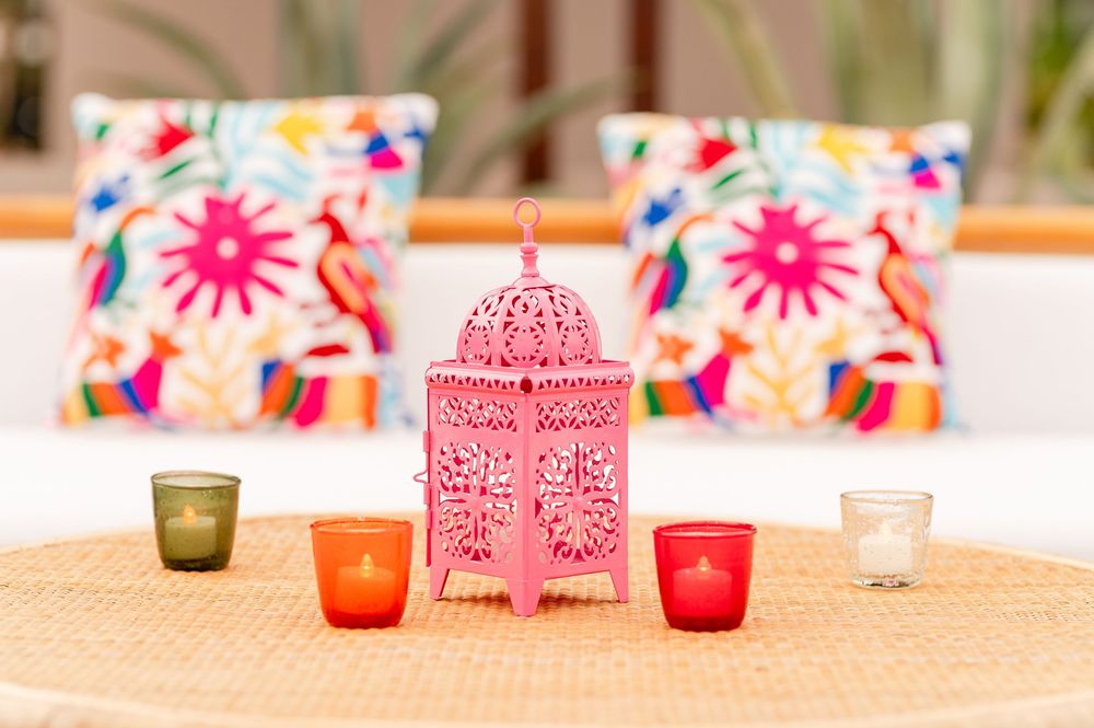 Pink Moroccan lantern with colorful votive candles on rattan table, vibrant embroidered pillows in background.
