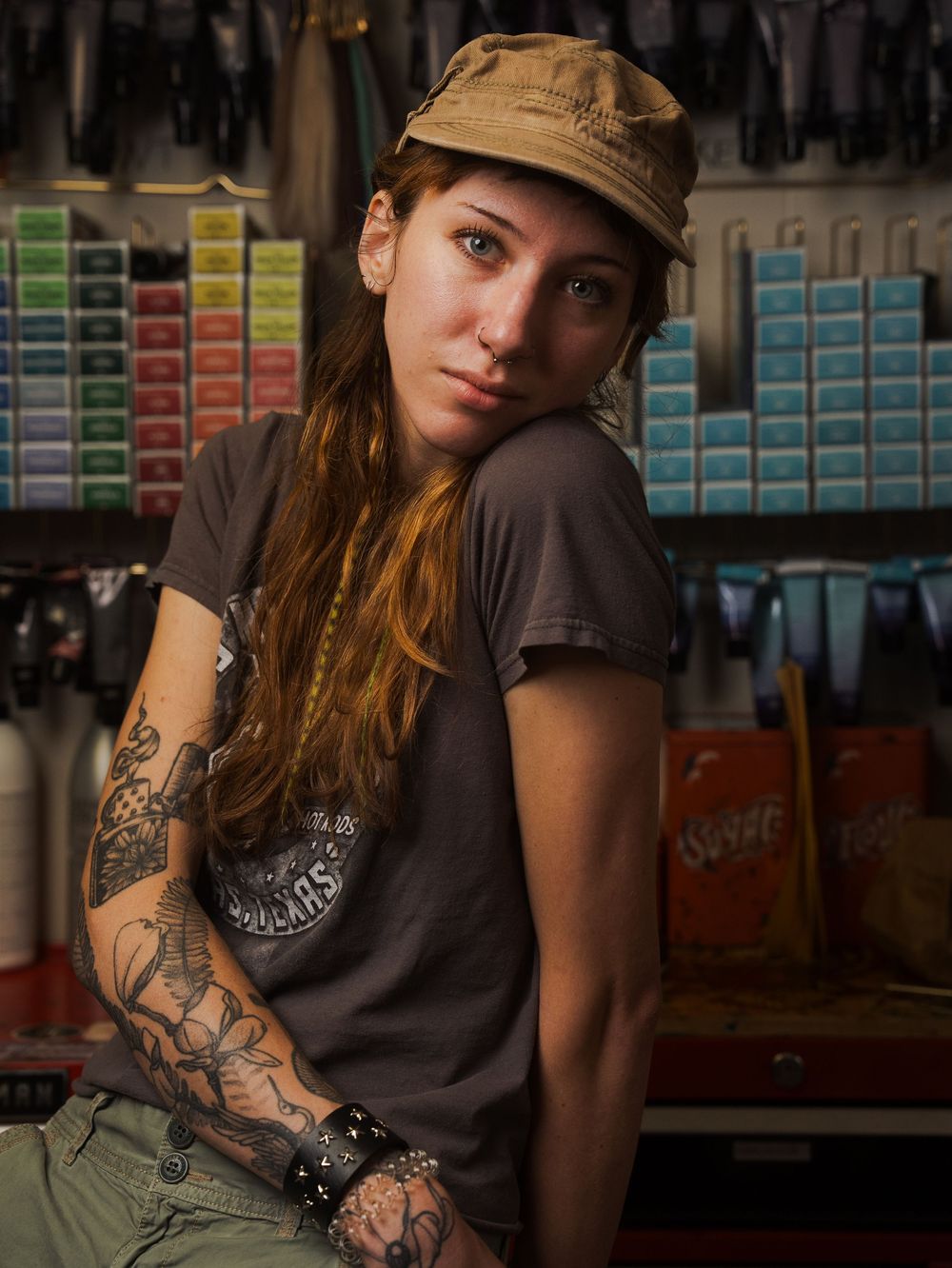 Tattooed woman in a cap and gray t-shirt poses in a shop with colorful product displays behind her.