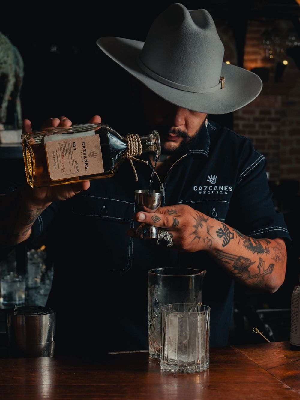 Bartender in a gray cowboy hat and Cazcanes shirt pours tequila from a bottle into a jigger above crystal glasses on a wooden bar.
