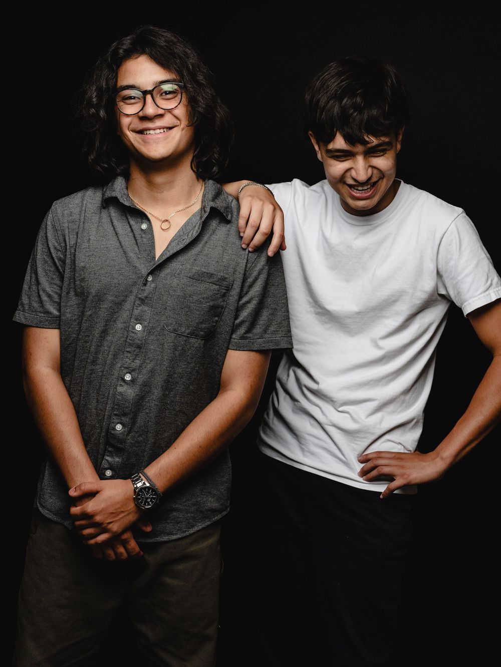 Two young men smiling and posing together against a dark background, one wearing glasses and a gray shirt, the other in a white tee.