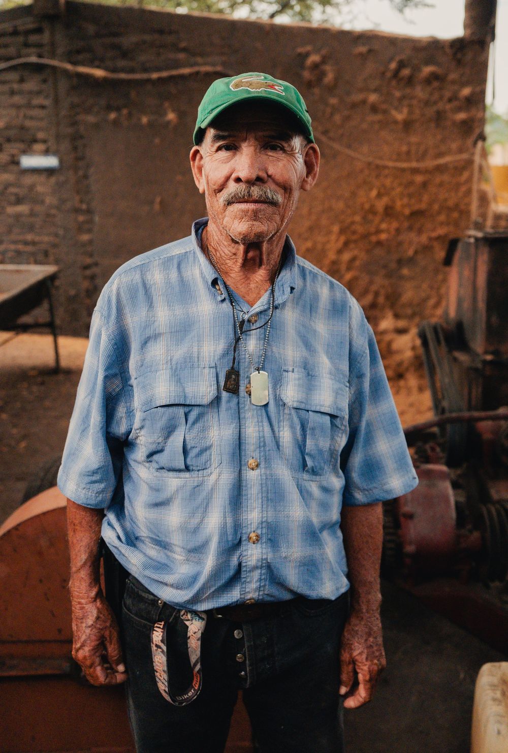 Elderly man in a green cap and blue plaid shirt stands outdoors beside rustic machinery and an adobe wall, wearing a dog tag necklace.