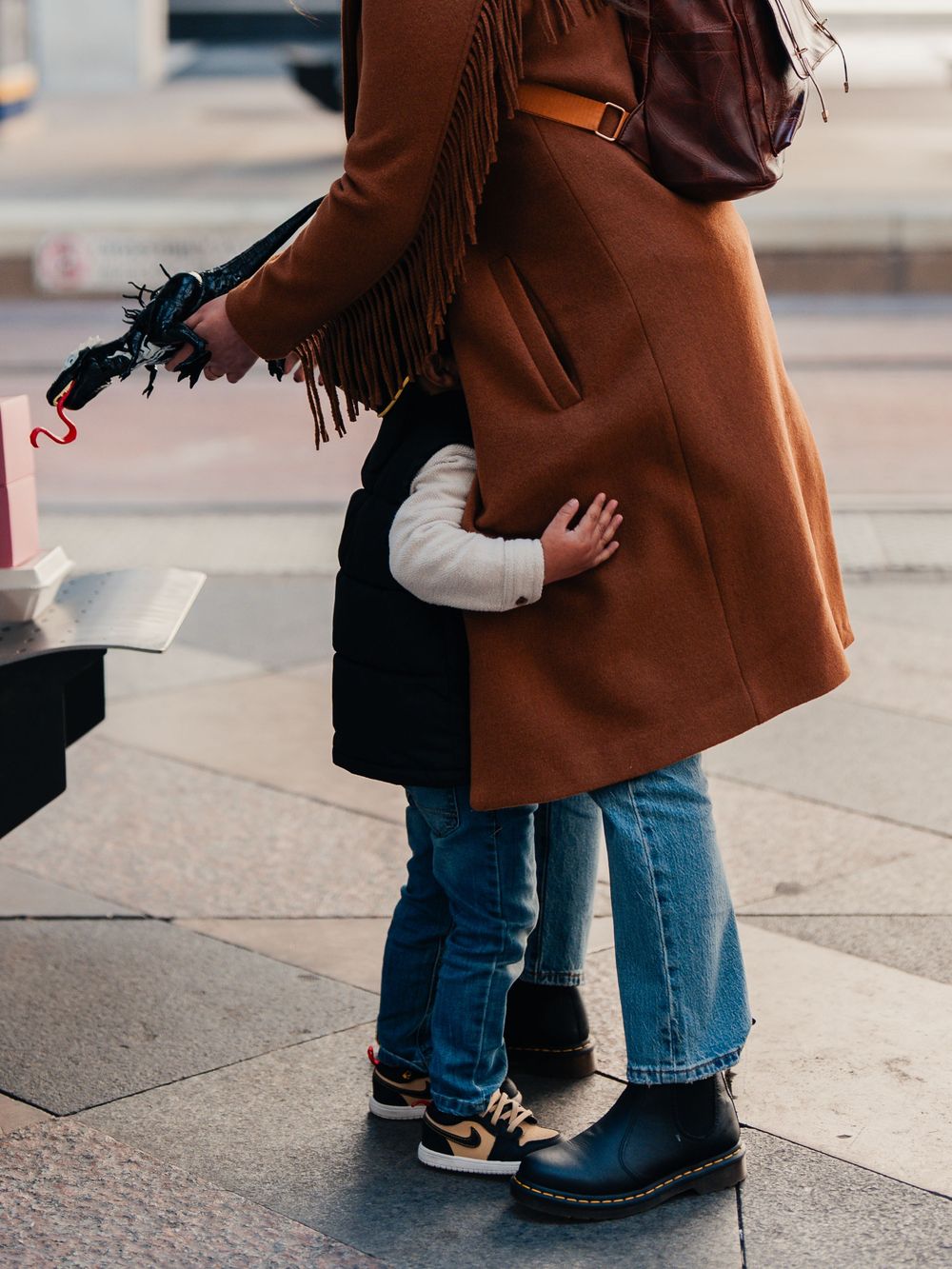 A mother in a brown coat and jeans hugs a small child on a city sidewalk, both wearing stylish boots.