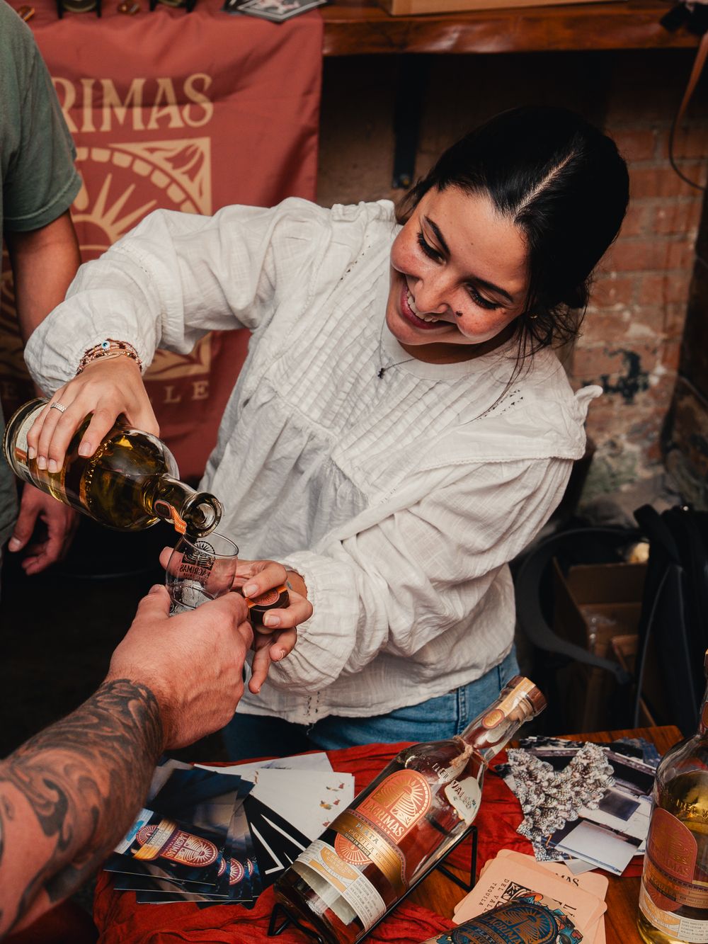 Woman in white sweater pouring whiskey at a tasting event with bottles on a red-draped table.