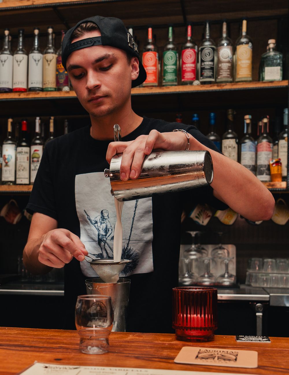 Bartender pouring cocktail through funnel strainer into glass at bar with bottles on shelves behind.