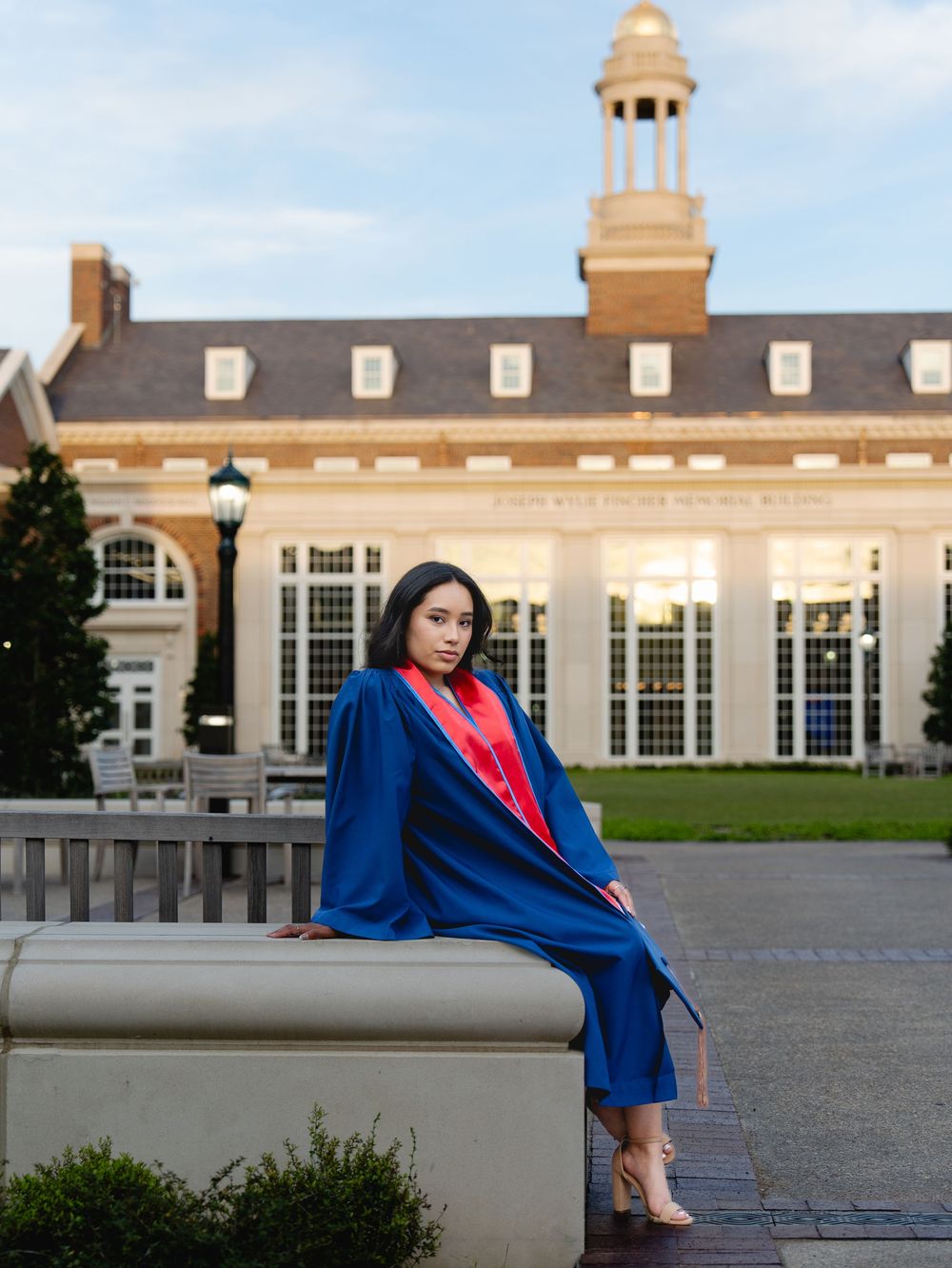 A young woman in blue graduation cap and gown poses on a stone bench in front of a classic university building.