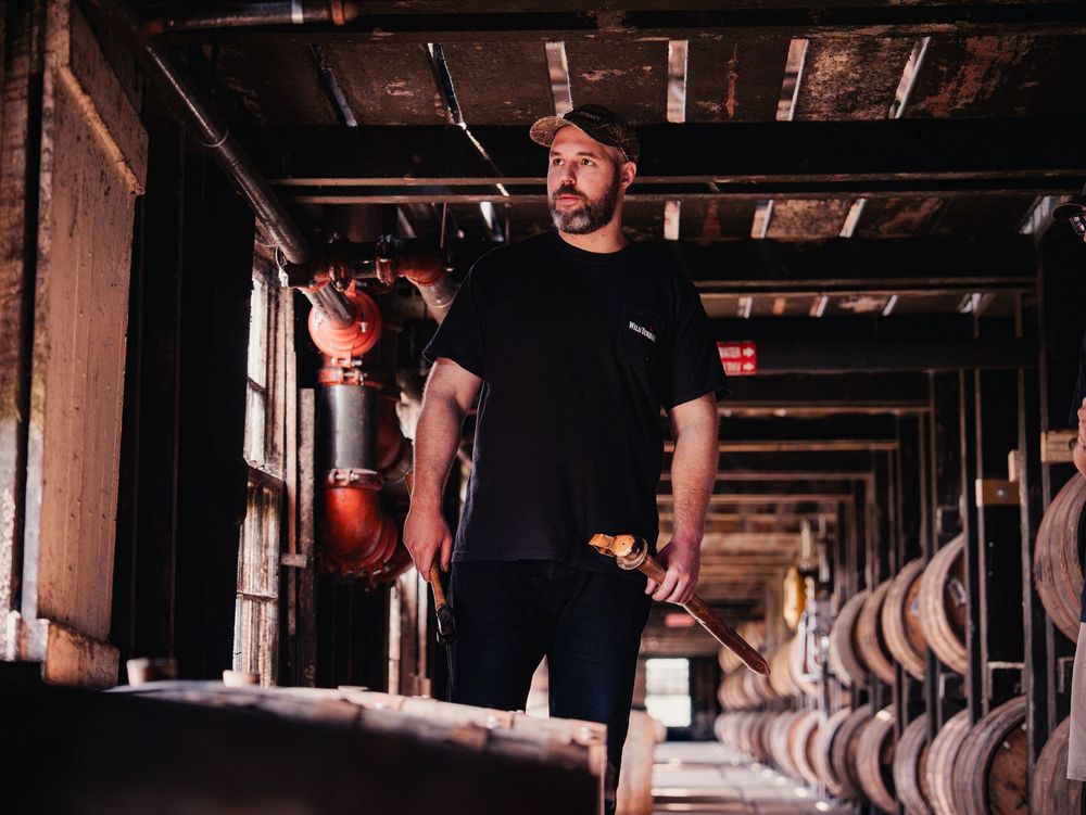 A man in a black t-shirt stands confidently among bourbon barrels in a dimly lit distillery warehouse.