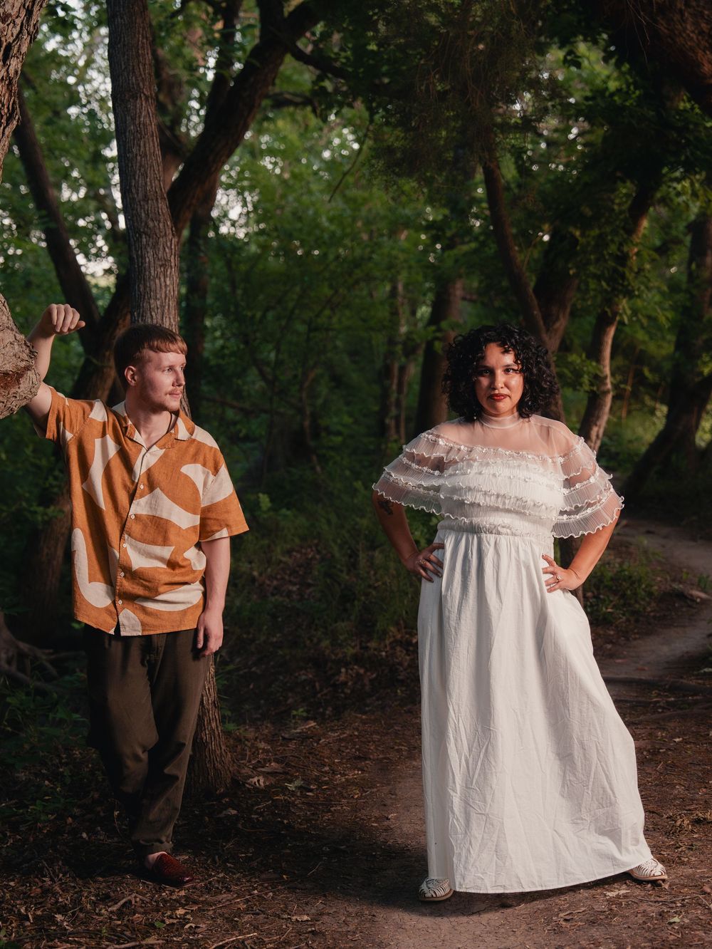 A couple poses in a forest, woman in white lace gown, man in brown patterned shirt, surrounded by trees.