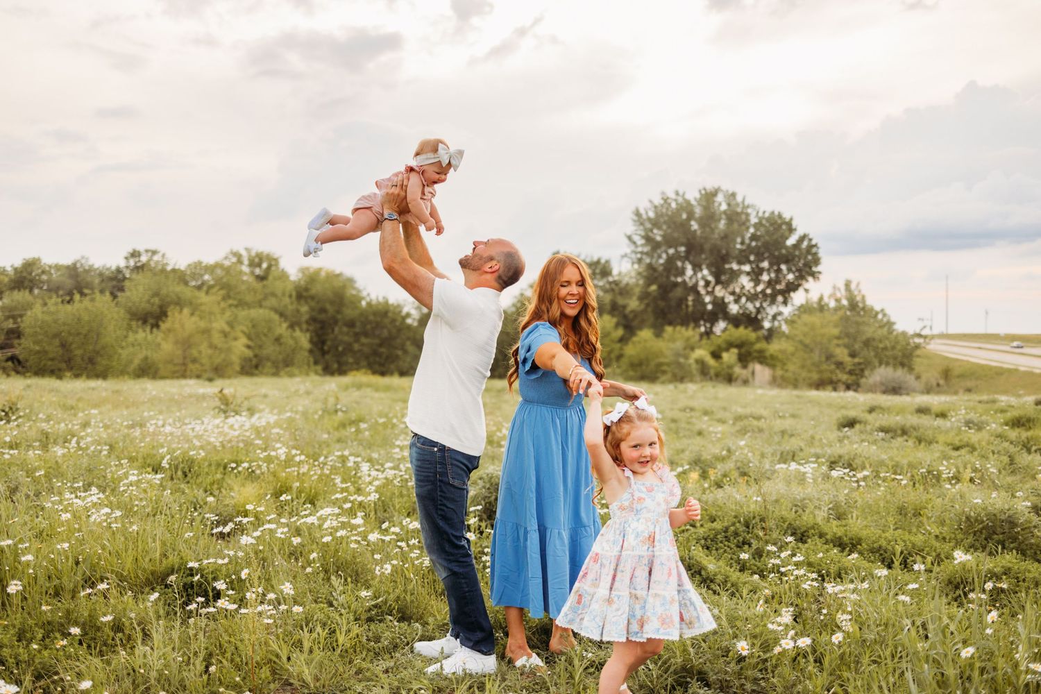 Happy family with two young daughters enjoying a sunny day in a blooming green meadow, father lifting baby girl in the air.