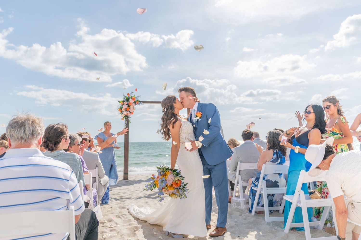Couple shares first kiss at romantic beach wedding ceremony with guests cheering under sunny skies.