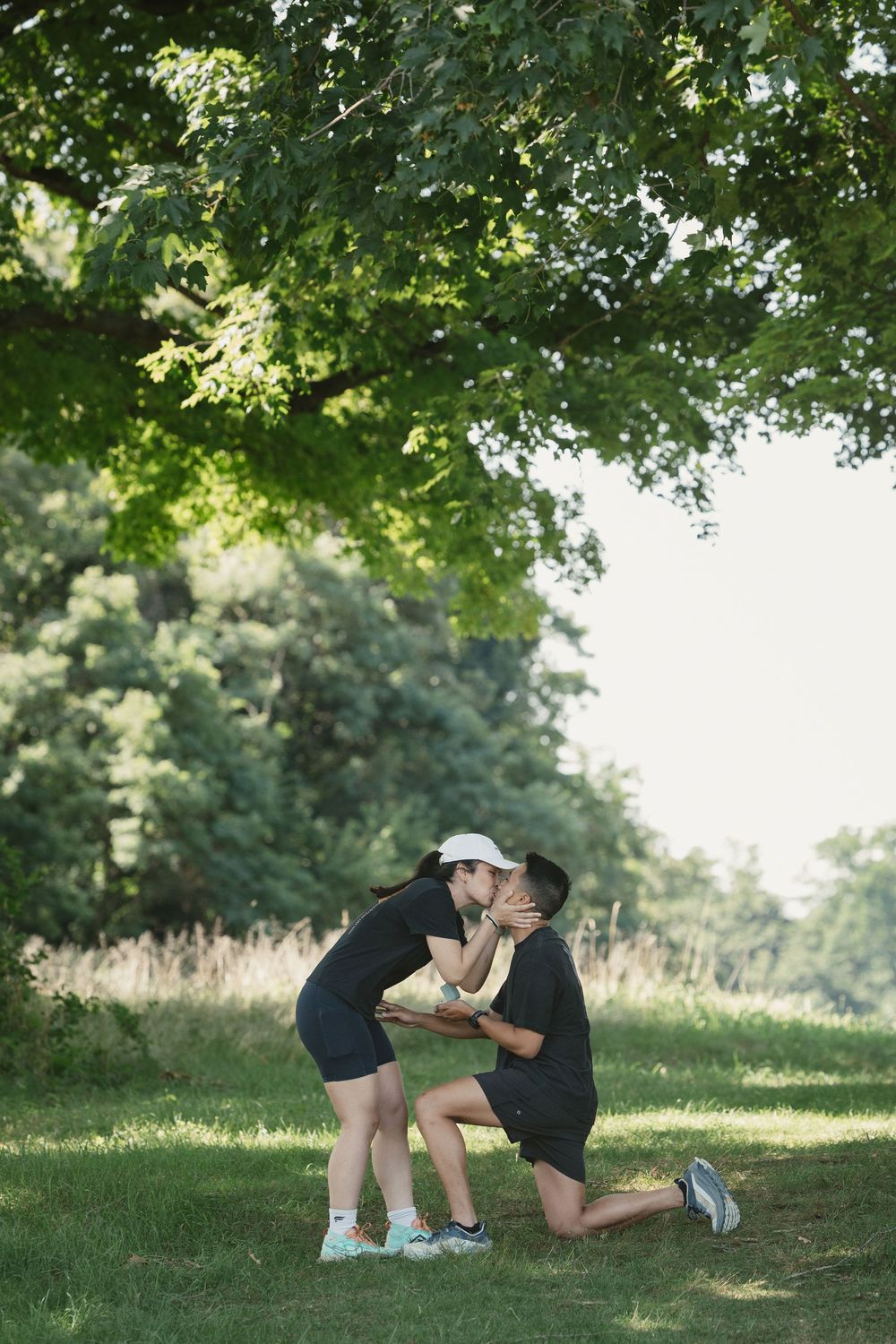 Couple sharing a romantic kiss in a lush green park, one partner kneeling on grass beneath large trees.