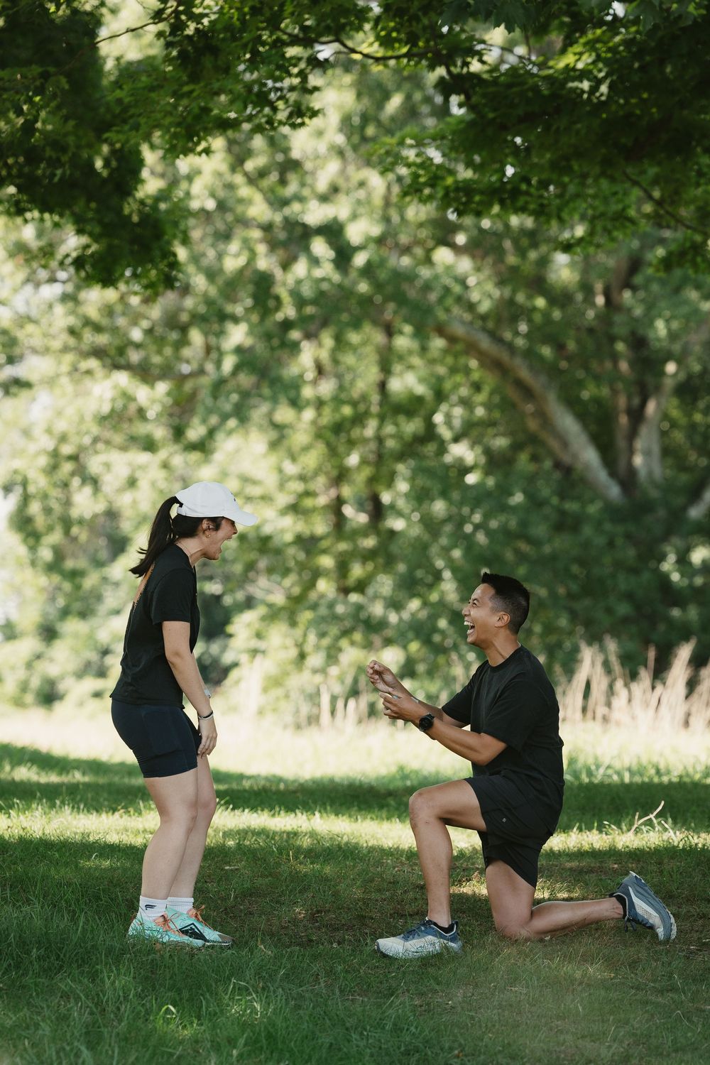 Man proposing on one knee to woman in black athletic wear during outdoor run in lush green park.