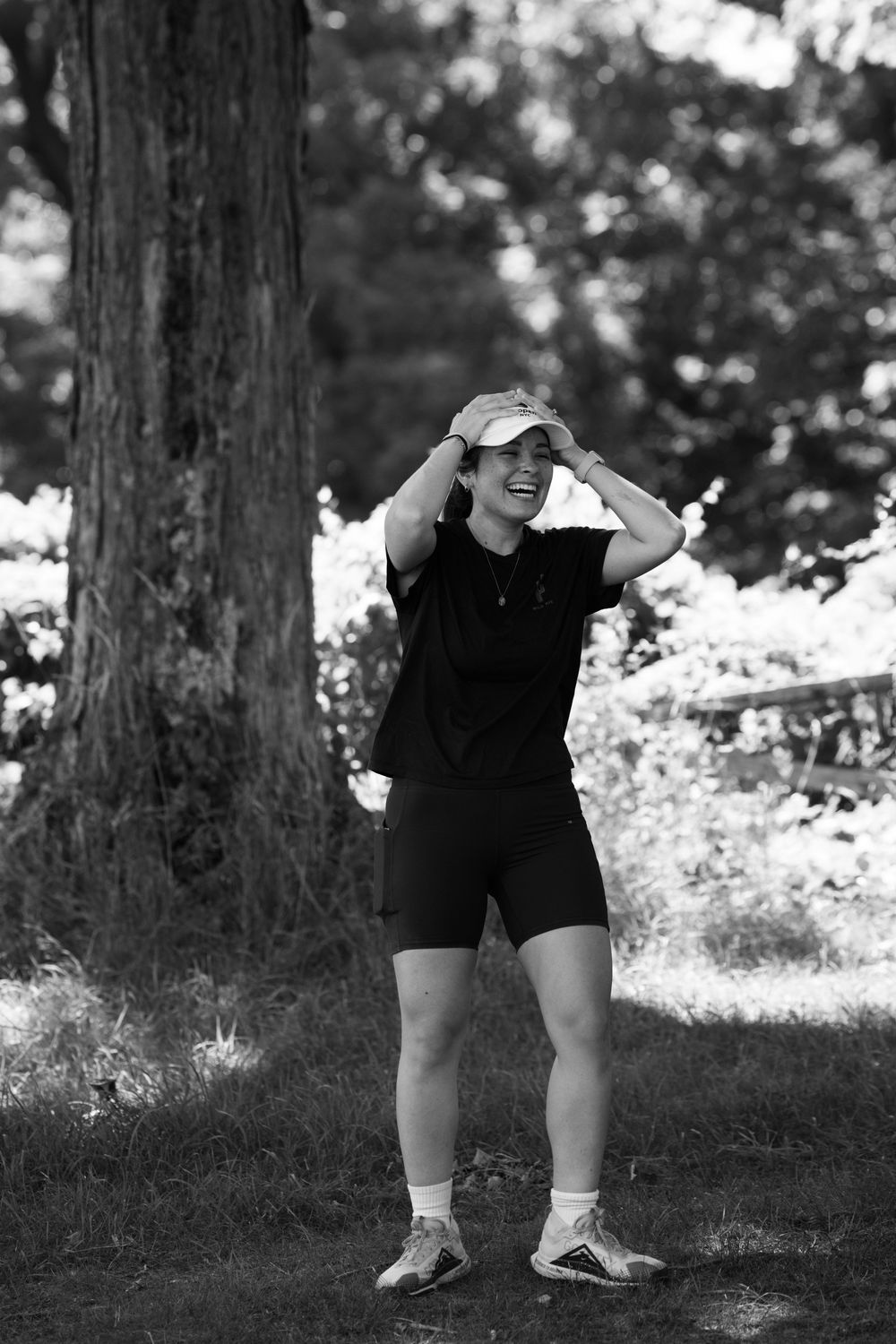Black and white photo of a smiling person in athletic wear adjusting their cap outdoors near a large tree.