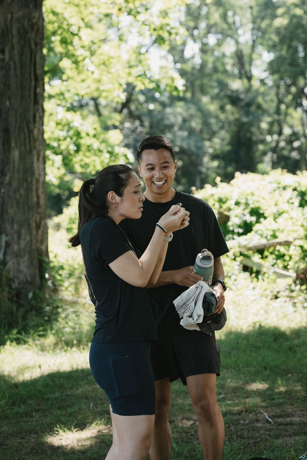 A laughing couple in black athletic wear taking a break outdoors, holding water bottles surrounded by green trees.