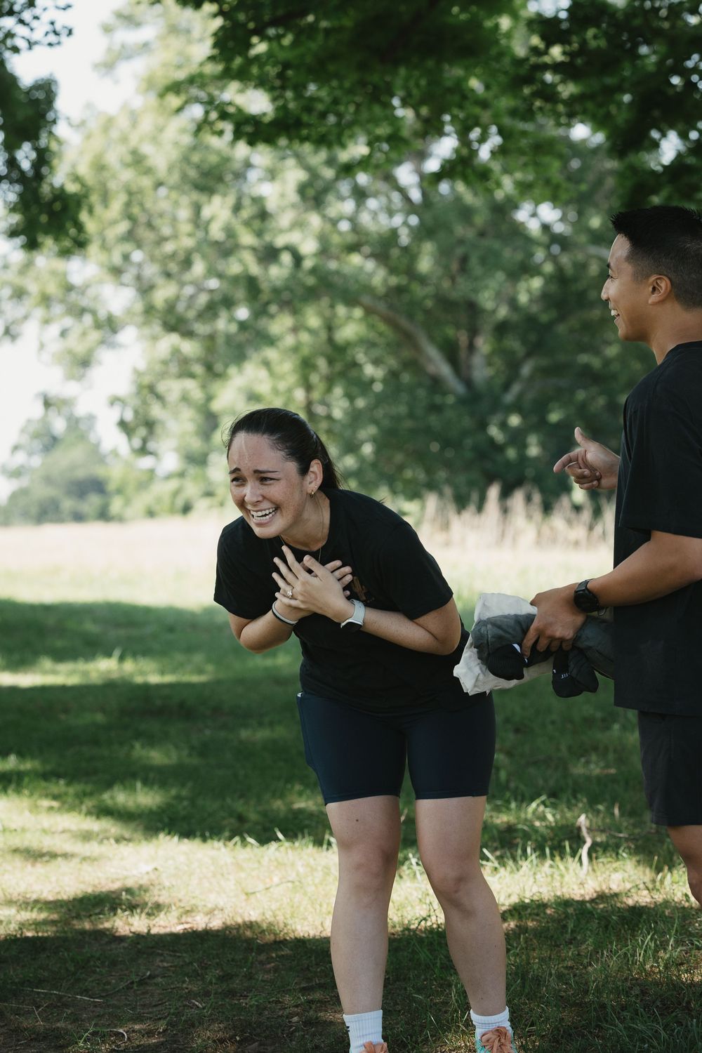 Two people in black activewear laughing together during an outdoor workout session in a sunny green park.