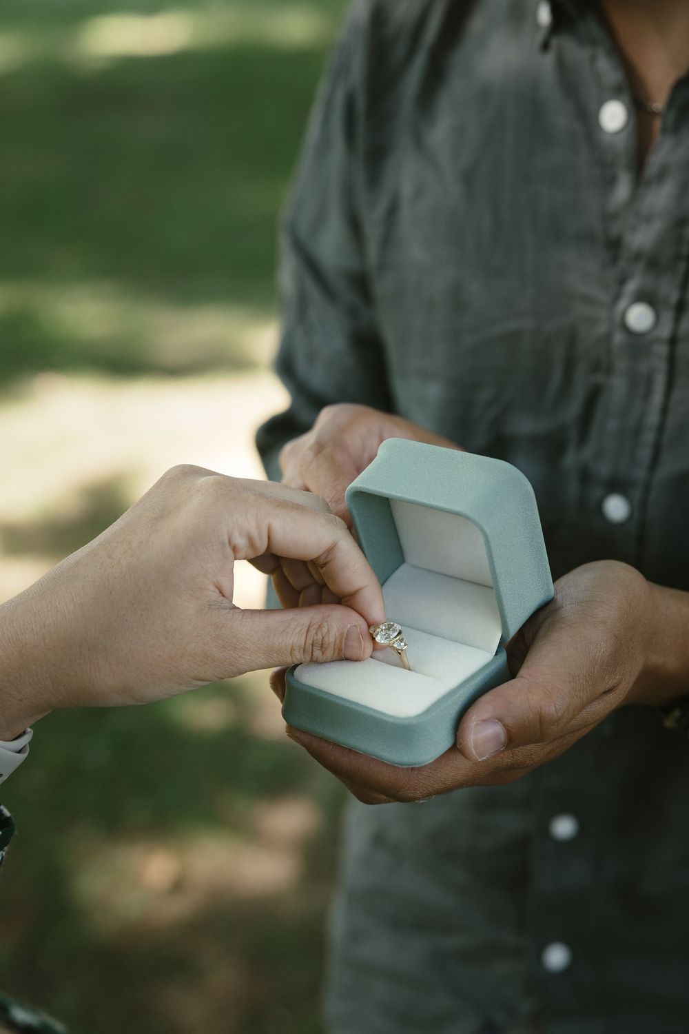 Person receiving a diamond engagement ring in a teal velvet box outdoors during a marriage proposal.